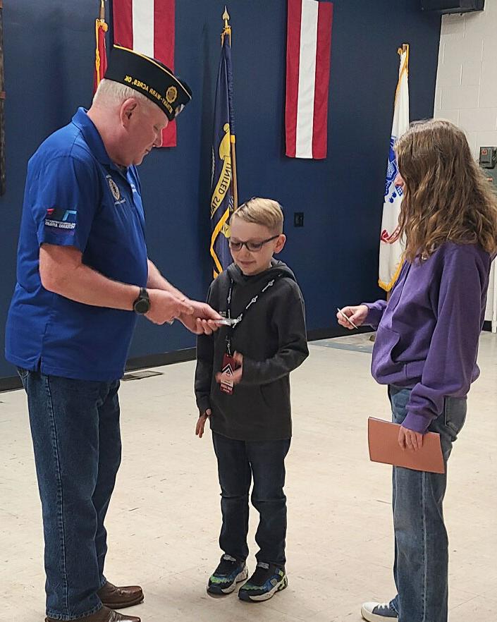 A man in a blue uniform and hat hands an item to a young boy, while a girl in a purple sweatshirt stands nearby holding a book. They are indoors with flags in the background.